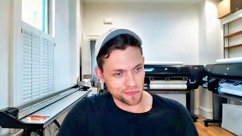 Young handsome guy in backwards cap smirks naughtily on webcam in print shop office.