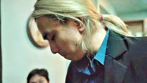 Blonde man with ponytail in suit gazes sideways with woman in background