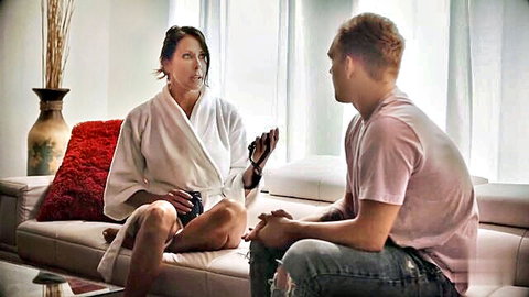 Brunette MILF in white robe exhales vape smoke while chatting with young guy on couch