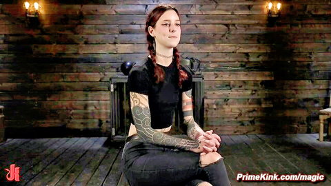 Tattooed redhead with pigtails sits smiling in BDSM dungeon ready for rough kinky domination and slave play