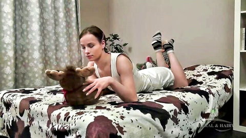 Petite brunette babe in short white dress cuddles stuffed reindeer toy on bed wearing socks