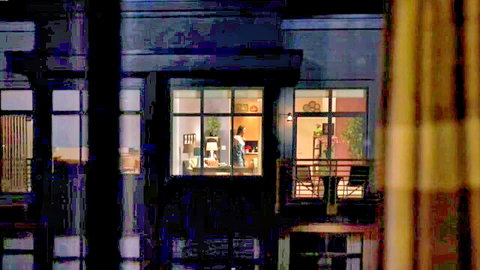 Voyeur view of sexy brunette in white blouse and skirt at desk in lit apartment window at night