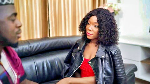 Busty black MILF in red cleavage top and leather jacket sits intimately with black stud on couch