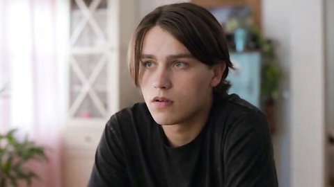 Handsome young man with tousled brown hair in black shirt gazes thoughtfully in sunlit bedroom