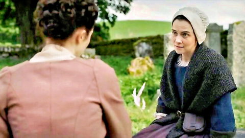 Two women in period dresses converse intimately near gravestones in lush green field