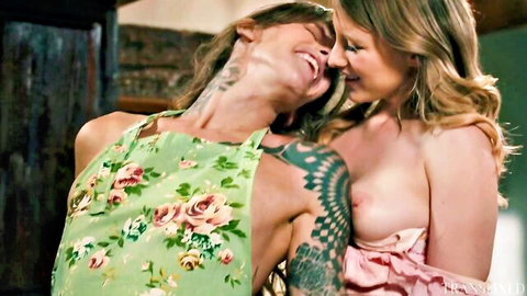 Tattooed blonde lesbian passionately kisses partner with medium tits exposed under green apron