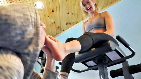 Blonde woman laughs as hooded guy sucks her bare toes on gym rower in foot fetish play