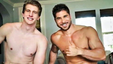 Two muscular shirtless gay hunks smiling and posing with shaka sign in bedroom