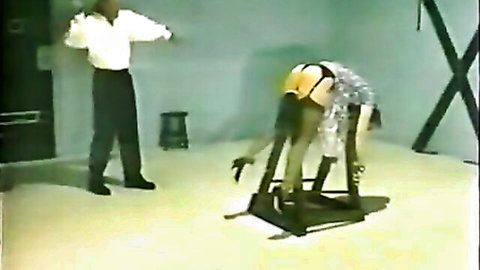 BDSM scene: restrained woman bent over, ass exposed, aggressive man preparing to whip