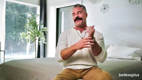 Mature gray-haired man with mustache sitting on bed gesturing in white sweater and beige pants