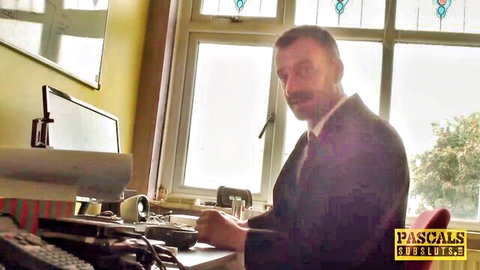 Suited man with mustache sits at desk in studio with monitors and window behind