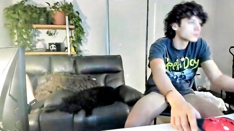 Curly-haired guy in Happy Day t-shirt sits on couch petting black cat in amateur webcam room