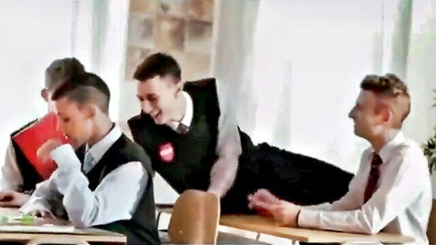 Schoolboys in uniforms smiling in classroom with one lying on desk hand near crotch