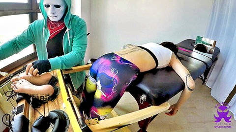 Masked man tickles woman's feet in stocks while she lies prone in galaxy leggings