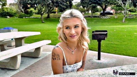 Blonde tattooed teen smiling with cleavage spilling from white camisole top at park picnic table