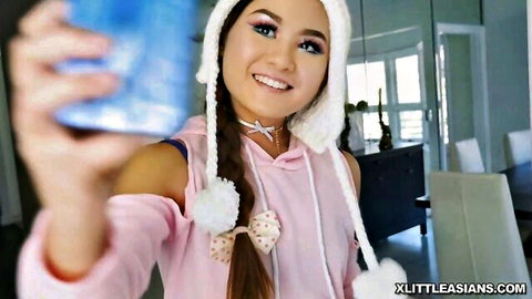 Petite Asian teen smiling selfie in pink off-shoulder hoodie fluffy hat holding blue cup