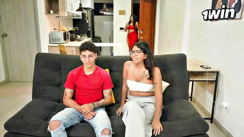 Curvy tattooed brunette in tight tube top and sweats cuddles boyfriend on couch with peeking woman in red dress