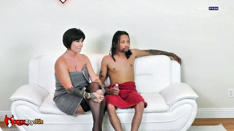 Tanned brunette MILF with big tits sits topless next to shirtless dreadlocked stud with tattoos on white couch