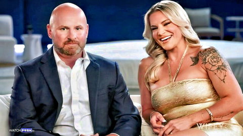 Smiling blonde tattooed wife in gold tube top with bald suited husband on couch