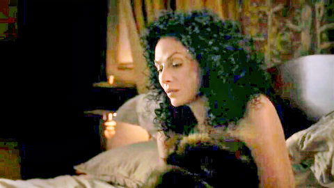 Curly-haired brunette with medium tits lying topless on bed in dimly lit bedroom