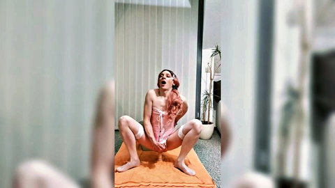 Redhead in pink lingerie squats wide-legged on orange mat holding dildo at her pussy