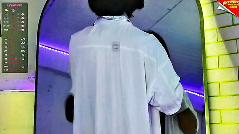Back view of long-haired guy in loose white shirt and pants posing in neon purple lit webcam room mirror