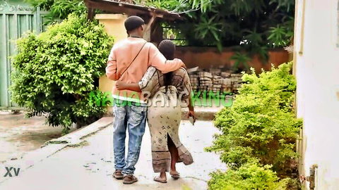 Sexy dark-skinned MILF in revealing dress embraced from behind by man outdoors