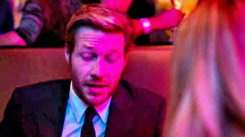 Suited blonde man with surprised expression in pink-lit club scene with nearby blonde woman