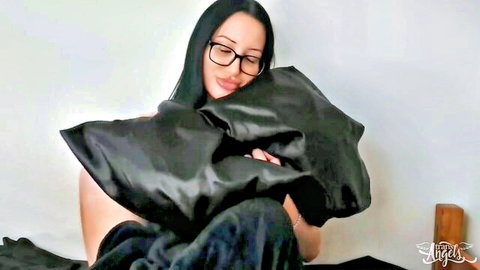 Busty ladyboy in glasses hugging black satin pillows, teasing solo pose