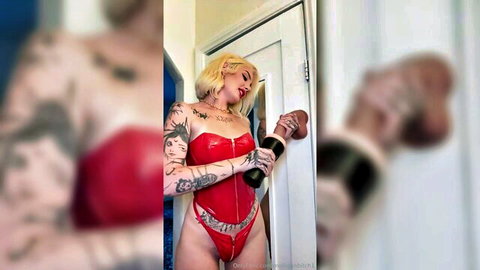 Tattooed blonde in shiny red latex corset holds massive black dildo teasingly against door