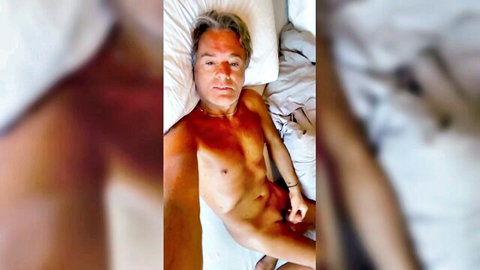 Nude muscular man with white hair lying on bed gripping his erect cock selfie
