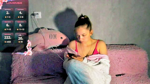 Curvy young brunette with big tits in pink bra on webcam holding phone with Lovense toys