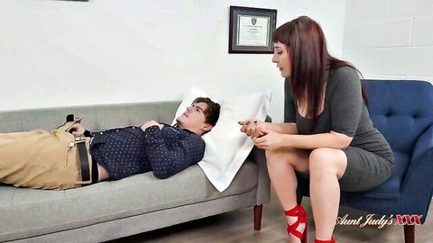 Brunette MILF therapist in gray dress leans over hypnotized young patient on couch