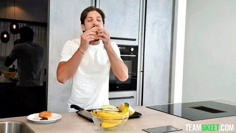 Man in white t-shirt bites juicy burger in modern kitchen with bananas and counter food