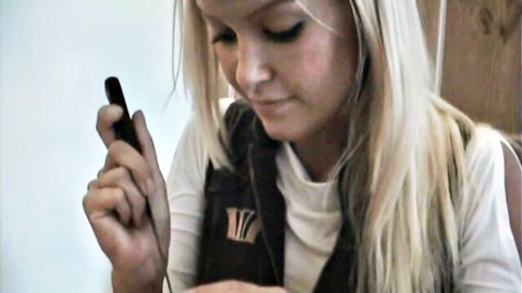 Blonde skinny girl in white tank top and vest holds phone smiling at camera