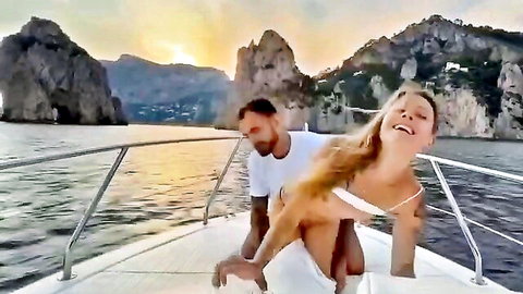 Blonde in white bikini smiles playfully leaning back on guy's lap on luxury boat at sunset Italian coast