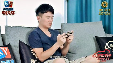 Handsome Asian man in blue V-neck shirt sits on gray couch holding smartphone casually