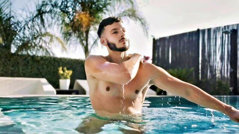 Athletic young brunette hunk posing naked in outdoor pool with water dripping on ripped abs