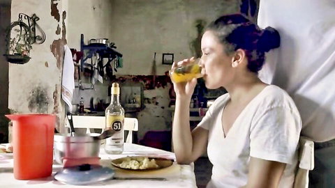Brunette woman with medium tits in low-cut white top sips orange juice at kitchen table showing cleavage