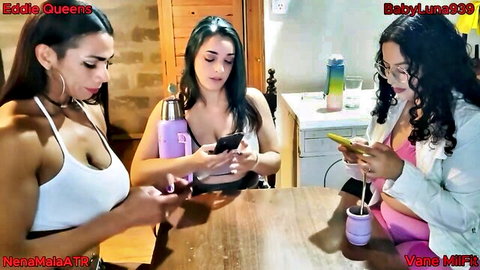 Three busty women in revealing tops scroll phones at kitchen table showing deep cleavage