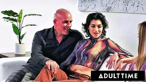 Bald man smiling with brunette in sheer dress and blonde woman in intimate threesome scene