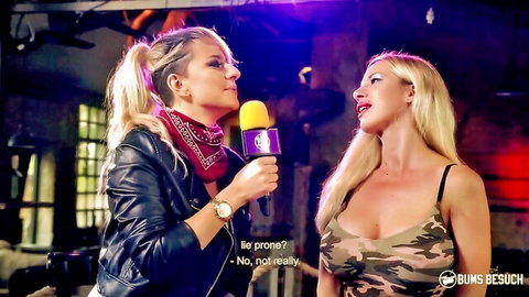 Busty blonde in camo top flashes deep cleavage during mic interview by leather-jacketed hottie in club