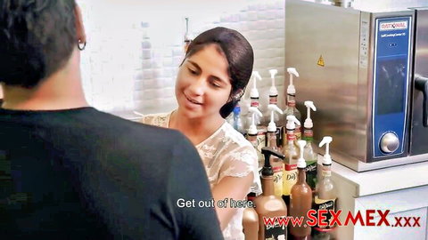 Smiling curvy Latina in lace top behind kitchen counter with condiment bottles facing black-clad guy in SexMex scene