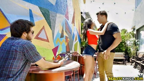 Hot Latina in tiny denim shorts embraced by muscular stud on colorful street while guy watches at cafe table