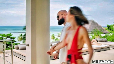 Brunette MILF in red sling bikini walks with BBC stud on tropical balcony in threesome preview