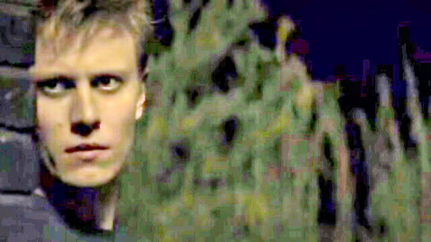 Intense blonde man with green eyes peeking from behind brick wall in dark night scene