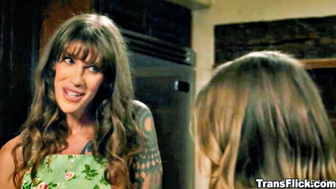Tattooed brunette trans woman in green floral dress smirks at young brunette neighbor in kitchen