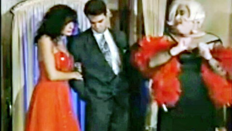 Brunette babe in sparkling red gown stands with suited man beside blonde drag queen in red feather boas