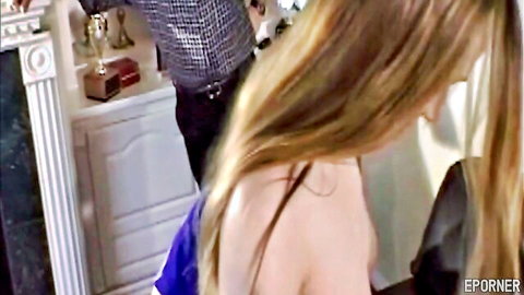 Blonde woman in blue top bends over with long hair as man stands behind her in retro room