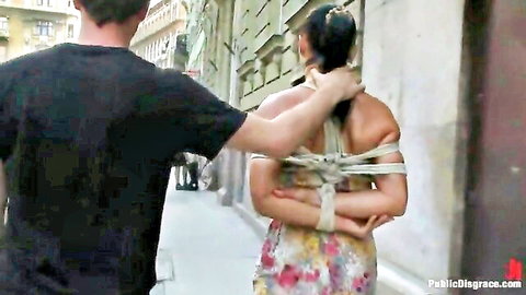 Man pulls ponytail of bound submissive woman in short floral dress on public street